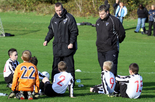 Mark Christie as a football coach with another coach talking to the young team